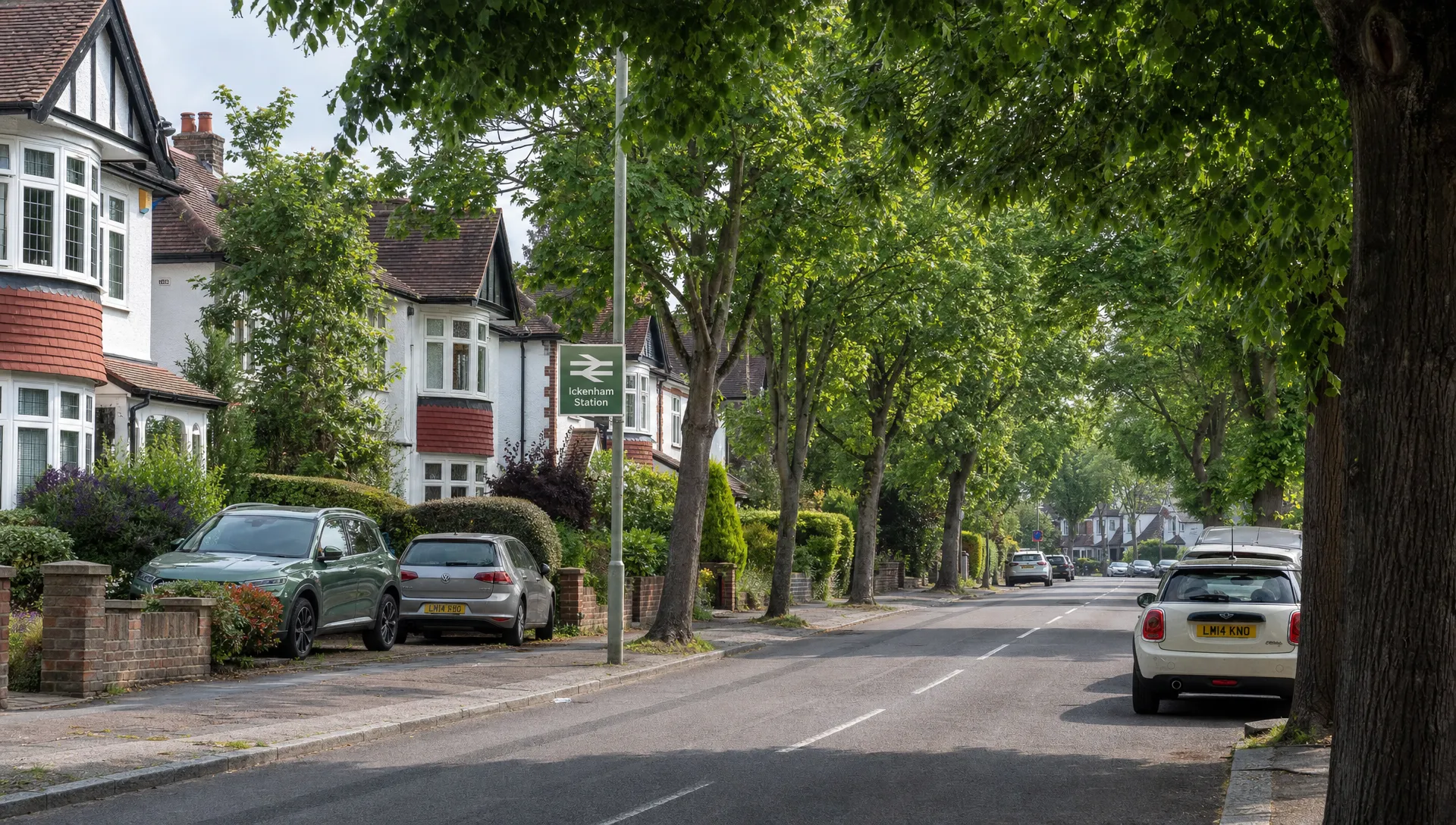 A wider view of a tree-lined residential road in Ickenham with 1930s semi-detached homes, front gardens, parked cars and a station sign in the distance, capturing the village setting and commuter appeal.