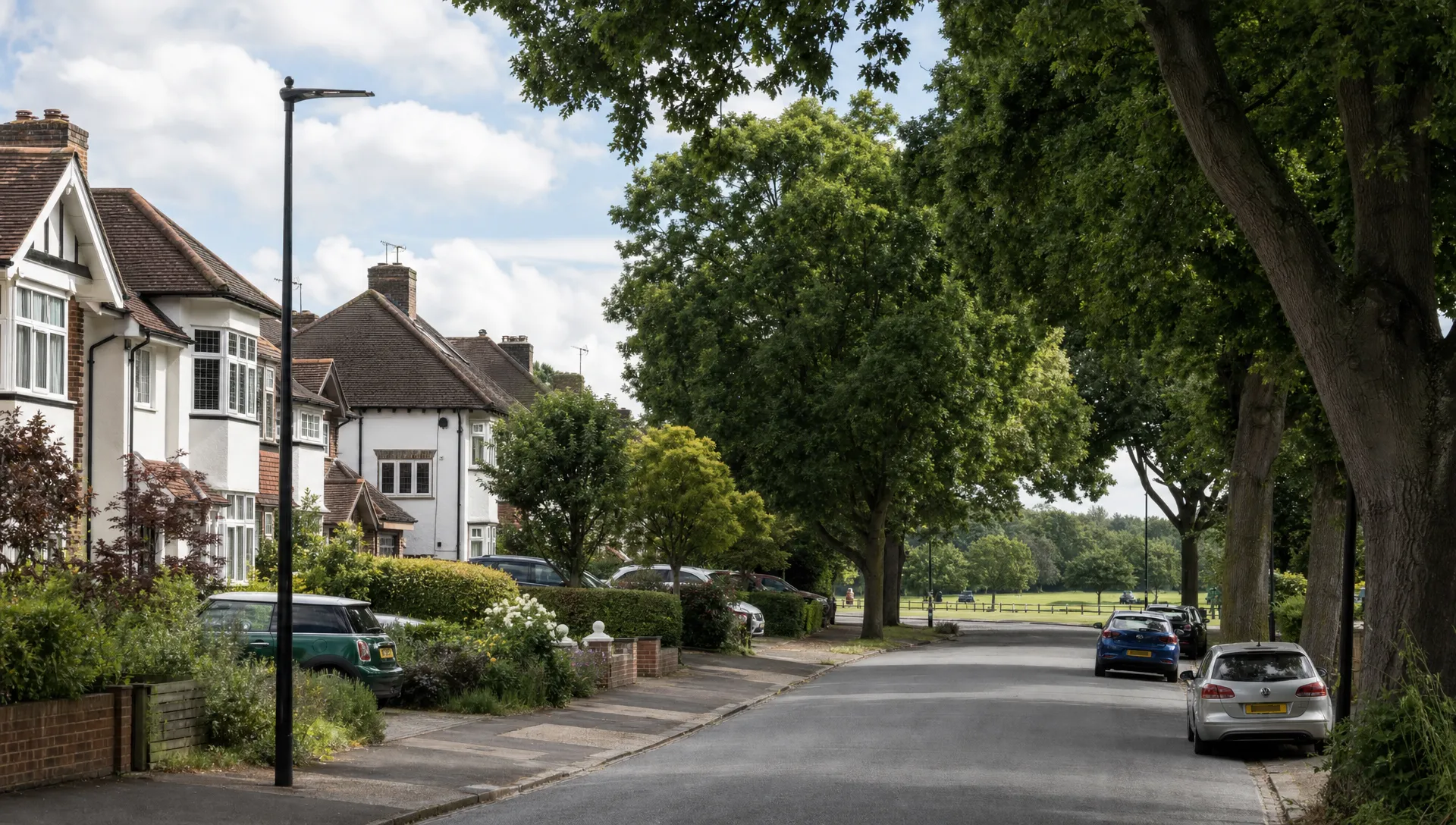 A residential Uxbridge street with 1930s semi-detached houses, mature front gardens, parked cars, leafy trees and a glimpse of a local park in the distance.